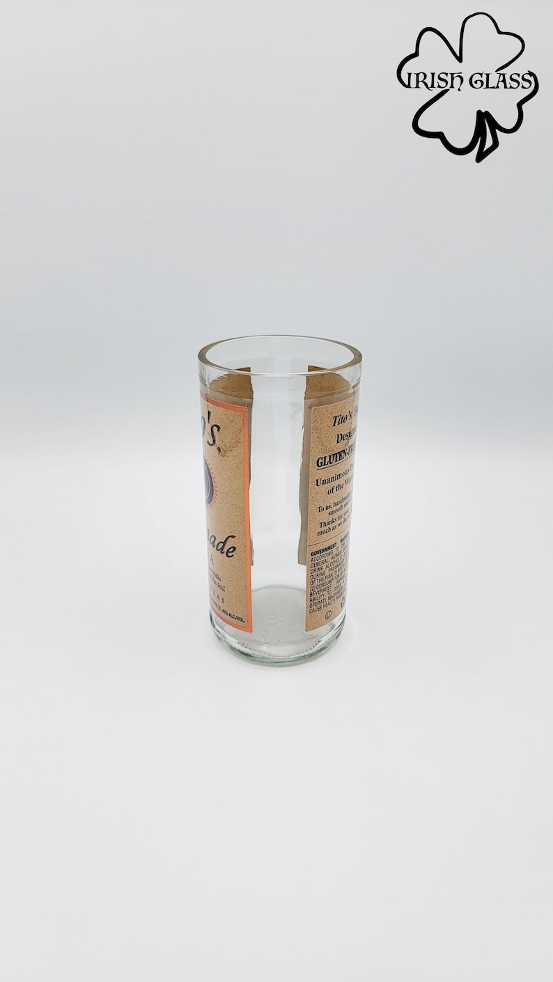 May include: A clear glass tumbler repurposed from a bottle, featuring a tan label with black text that reads Tito's Handmade Vodka, Gluten-Free, and includes a government warning. The top rim is smooth and the base is circular and transparent.