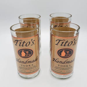 May include: A set of four clear glass tumblers, each featuring a tan label with the text Tito's Handmade Vodka, an illustration of a pot still, and Austin, Texas. The glasses are arranged in a square formation against a plain white background.