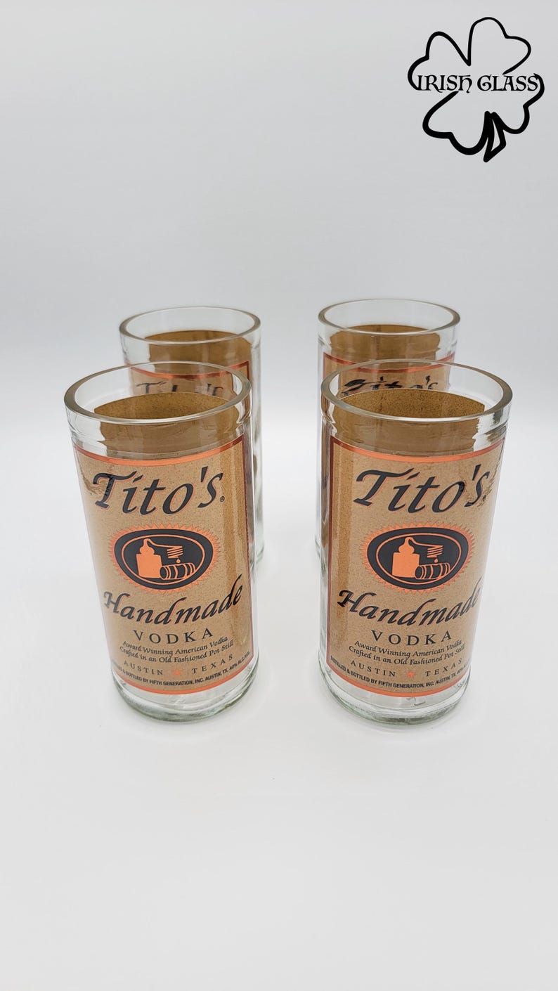 May include: A set of four clear glass tumblers, each featuring a tan label with the text Tito's Handmade Vodka, an illustration of a pot still, and Austin, Texas. The glasses are arranged in a square formation against a plain white background.