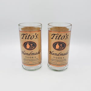 May include: Two clear glass tumblers featuring the Tito's Handmade Vodka label in tan with black text and a copper-colored still icon. The label reads Tito's Handmade Vodka, Austin, Texas. An Irish Glass logo is visible in the top right corner.