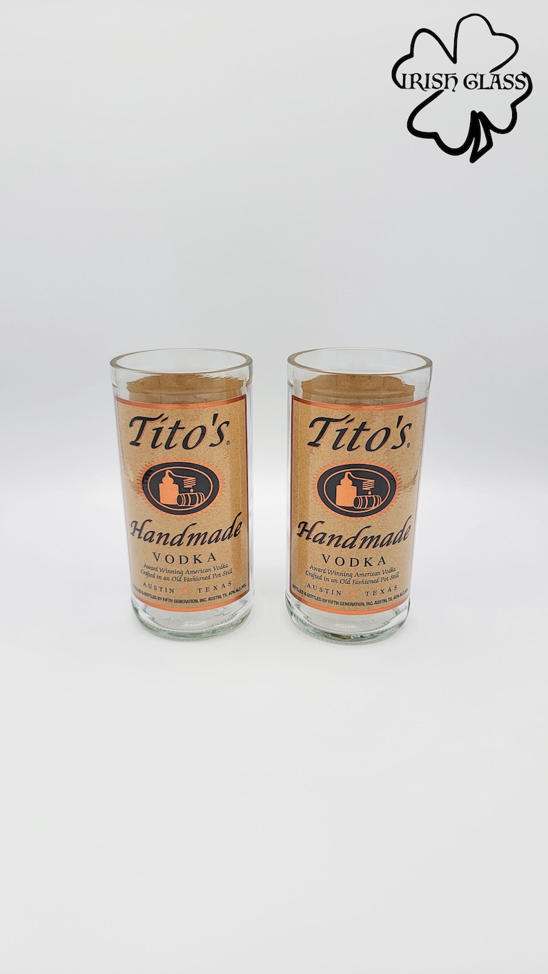 May include: Two clear glass tumblers featuring the Tito's Handmade Vodka label in tan with black text and a copper-colored still icon. The label reads Tito's Handmade Vodka, Austin, Texas. An Irish Glass logo is visible in the top right corner.