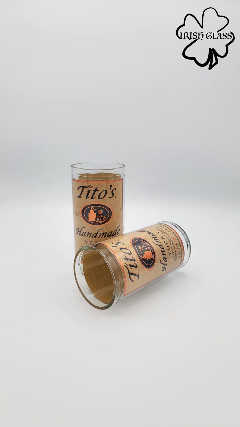 May include: Two clear glass tumblers featuring the Tito's Handmade Vodka label with tan paper and black text. One glass stands upright while the other rests horizontally in front. The logo includes a still illustration and the text Tito's Handmade.