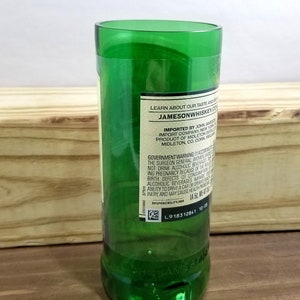 May include: A green glass bottle with a label that reads "LEARN ABOUT OUR TASTE AND STYLE" and "JAMESONWHISKEY.CO". The label also includes information about the importer, product, and government warnings. The bottle is sitting on a wooden surface.