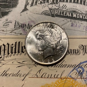 Puede incluir: Una moneda de plata Peace Dollar con el a&ntilde;o 1922 inscrito en la parte inferior. La moneda presenta un perfil de una mujer con el pelo suelto y las palabras "In God We Trust" inscritas en la parte inferior.