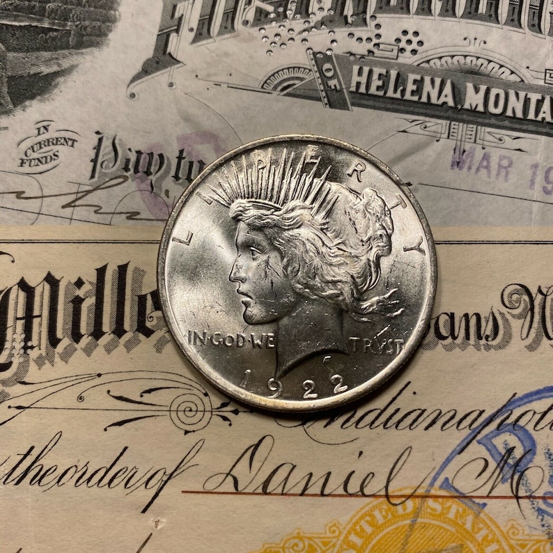 Puede incluir: Una moneda de plata Peace Dollar con el a&ntilde;o 1922 inscrito en la parte inferior. La moneda presenta un perfil de una mujer con el pelo suelto y las palabras "In God We Trust" inscritas en la parte inferior.