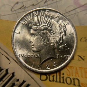 Puede incluir: Una moneda de plata Peace Dollar con el a&ntilde;o 1924 inscrito en la parte inferior. La moneda presenta el perfil de una mujer con una corona de rayos detr&aacute;s de su cabeza. La inscripci&oacute;n "In God We Trust" es visible en la parte inferior de la moneda.