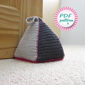 May include: A gray and black crocheted doorstop in the shape of a pyramid with a pink trim. The doorstop is sitting on a light beige carpet. The text "PDF pattern x" is in a pink circle with white stripes.