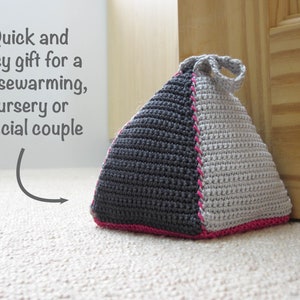 May include: A gray and white crocheted doorstop in the shape of a pyramid with a pink trim. The doorstop has a loop handle.