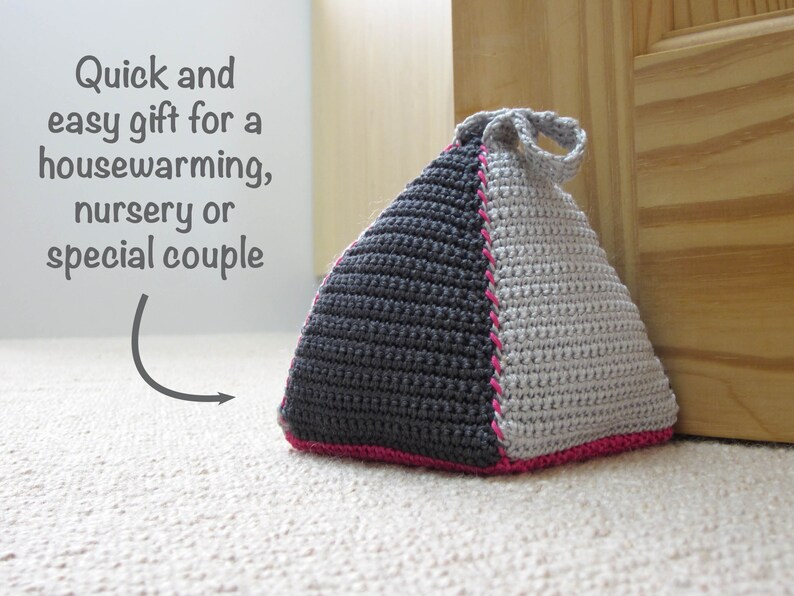 May include: A gray and white crocheted doorstop in the shape of a pyramid with a pink trim. The doorstop has a loop handle.