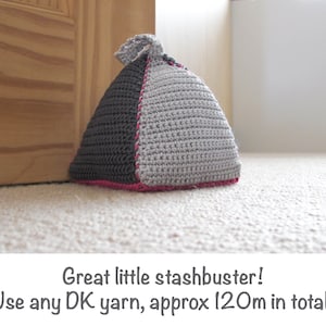 May include: A gray and black crocheted doorstop in the shape of a triangle with a pink trim.  Great little stashbuster! Use any DK yarn, approx 120m in total.