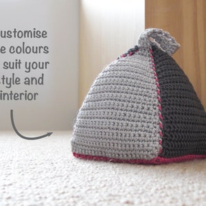 May include: A gray and black crocheted doorstop with a pink trim. The doorstop is shaped like a triangle and has a knotted top.