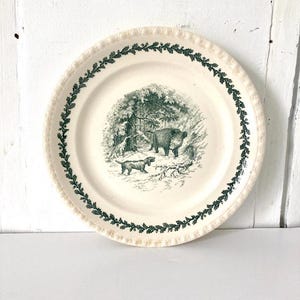 May include: A cream-colored ceramic plate featuring a dark green transferware illustration of a wild boar and a hunting dog in a forest setting. The rim is decorated with a repeating dark green oak leaf and acorn pattern and a raised beaded edge.
