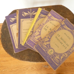 May include: A stack of vintage French paperback books with purple covers and yellow spines. The titles include "Le Médecin Malgré Lui" by Molière, "Le Barbier de Séville" by Beaumarchais, and "Fleurus" by Baudel. The books are part of the "Classiques Larousse" series.