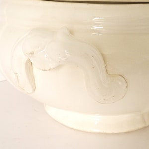 May include: White ceramic soup tureen with ornate handles. The tureen has a smooth, glossy finish and a classic design.
