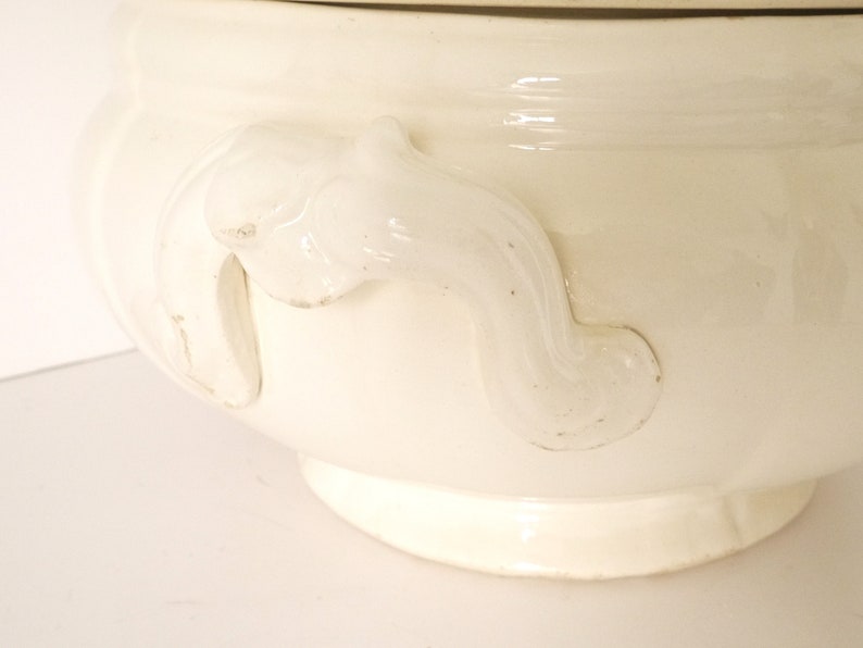 May include: White ceramic soup tureen with ornate handles. The tureen has a smooth, glossy finish and a classic design.