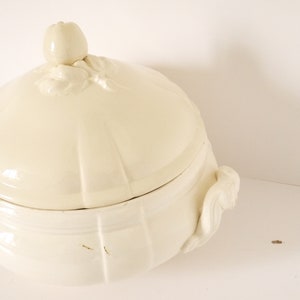 May include: A white ceramic soup tureen with a lid and two handles. The lid has a decorative knob in the shape of an apple.