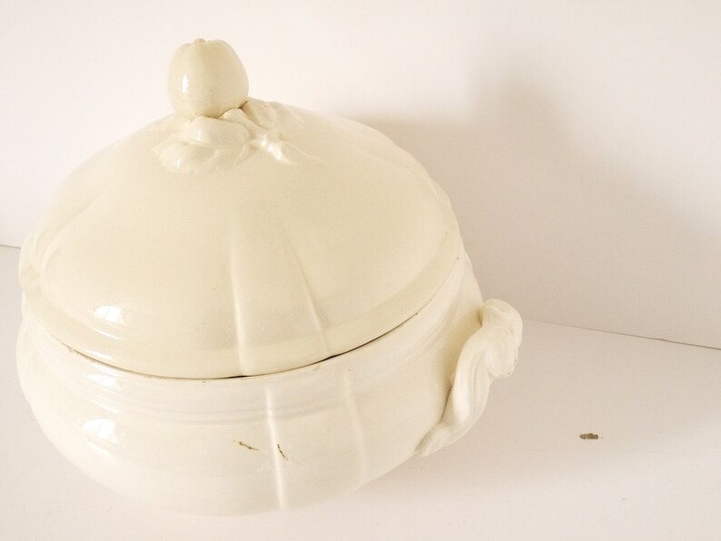 May include: A white ceramic soup tureen with a lid and two handles. The lid has a decorative knob in the shape of an apple.