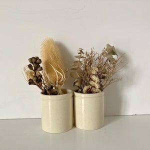 May include: Two cylindrical off-white ceramic crocks, each containing a dried floral arrangement. The left crock holds brown seed pods and a tall, spiky tan teasel. The right crock features a mix of dried eucalyptus leaves and light-colored grasses.