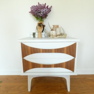 May include: White nightstand with two drawers and brown wood trim. The drawers have a curved design. The nightstand has four legs.
