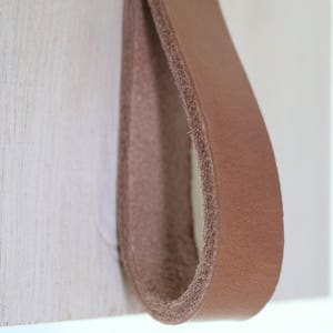 Peut inclure: Une poignée en cuir marron en forme de boucle avec une vis en métal doré. La poignée est fixée à une surface en bois.
