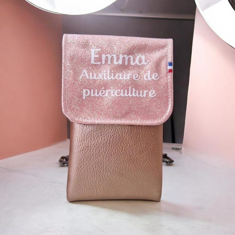 May include: A pink and gold purse with the name "Emma" and the text "Auxiliaire de pu&eacute;riculture" in white lettering. The purse has a flap closure and a small French flag emblem.