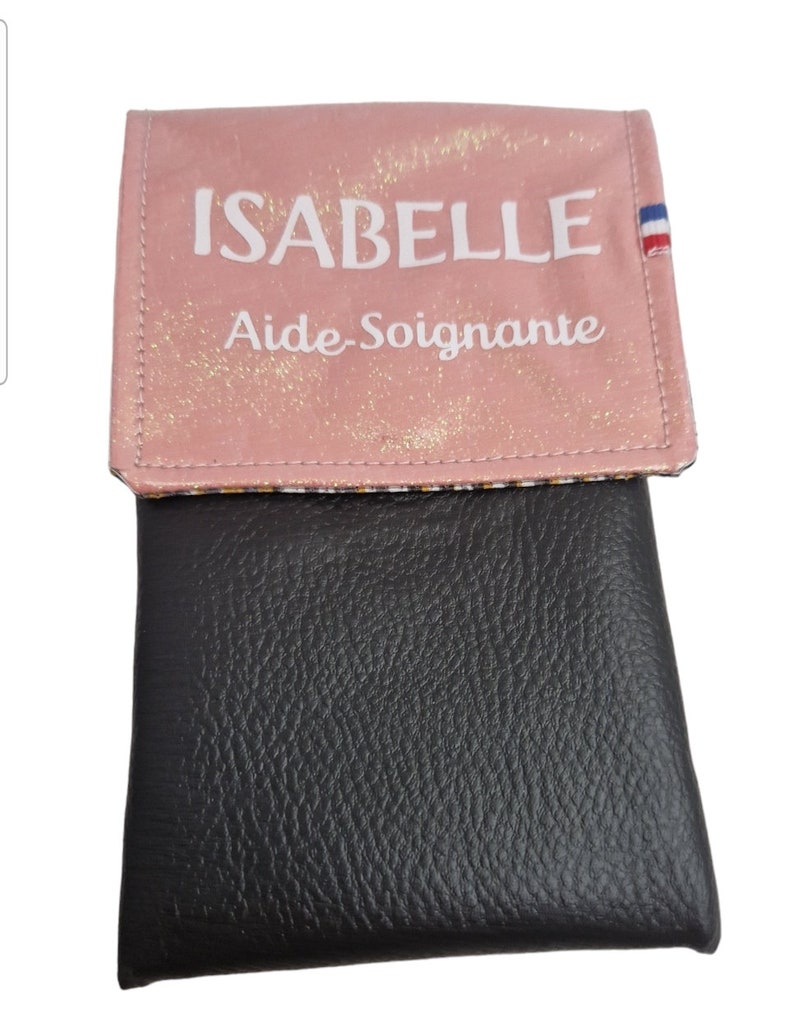 May include: A pink and black wallet with the name "ISABELLE" and the words "Aide-Soignante" printed on the front. The pink flap has a glittery texture and a small French flag detail. The bottom portion is black.
