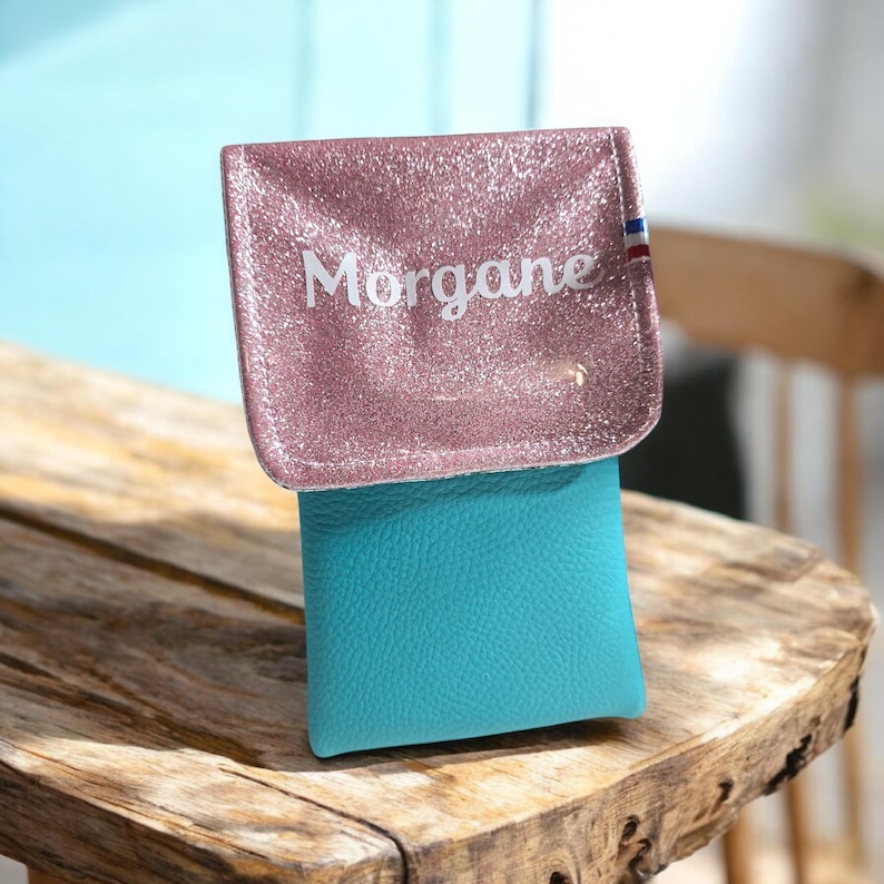 May include: A small, two-toned pouch with a pink glittery flap and a turquoise body. The flap is embossed with the name "Morgane" in white lettering. A small tricolor detail is visible on the right side of the flap. The pouch is sitting on a wooden surface.
