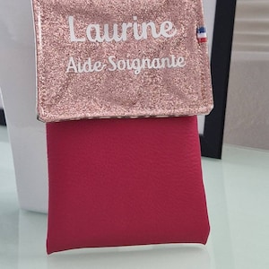 May include: A pink glittery wallet with the name "Laurine" and the words "Aide Soignante" printed in white. The wallet has a red lower section and a small French flag emblem.