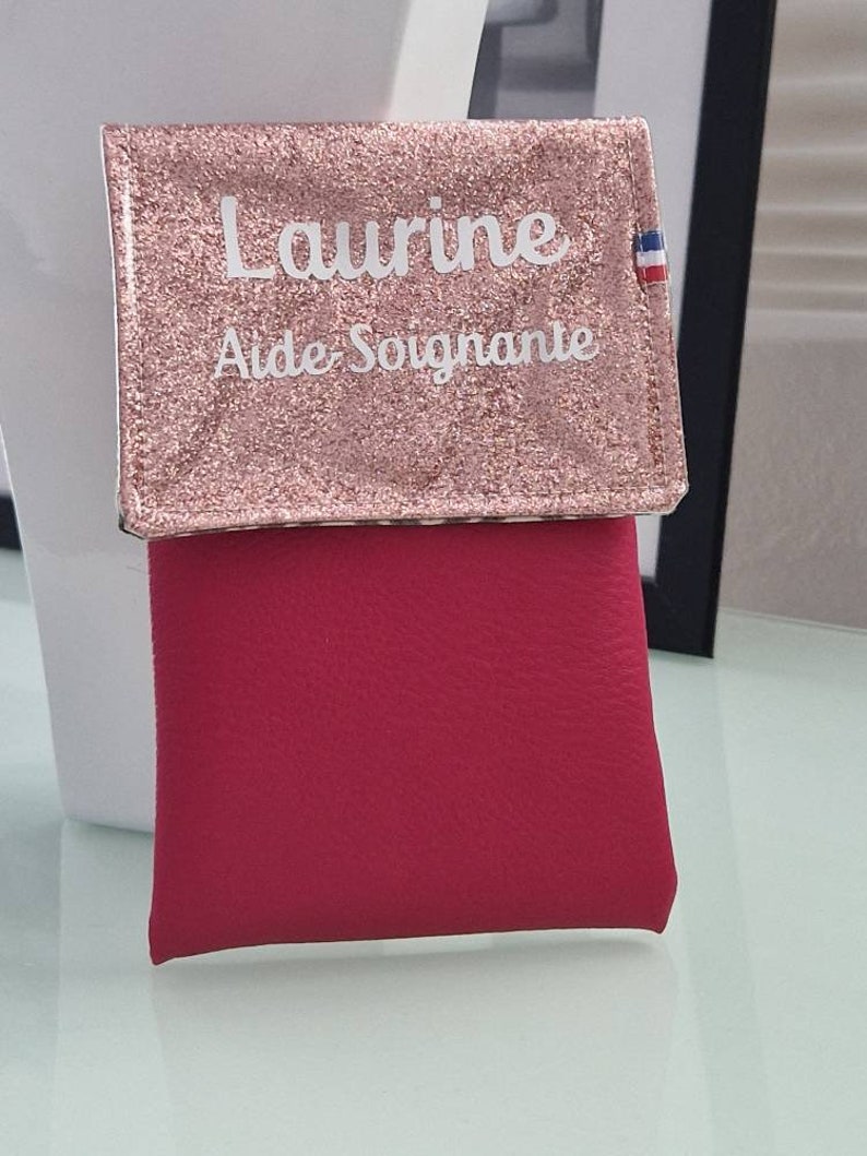 May include: A pink glittery wallet with the name "Laurine" and the words "Aide Soignante" printed in white. The wallet has a red lower section and a small French flag emblem.