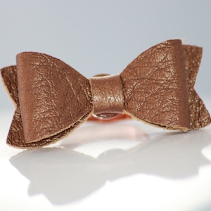 Peut inclure: Un nœud papillon en cuir marron, probablement un accessoire pour cheveux, est présenté sur un fond blanc. Le nœud a une surface texturée et une forme classique de nœud papillon. L'accessoire est conçu pour être porté.