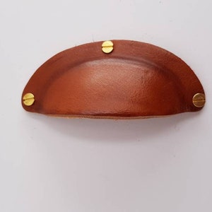 Può includere: Una maniglia per cassetti in pelle marrone con quattro viti in metallo dorato.