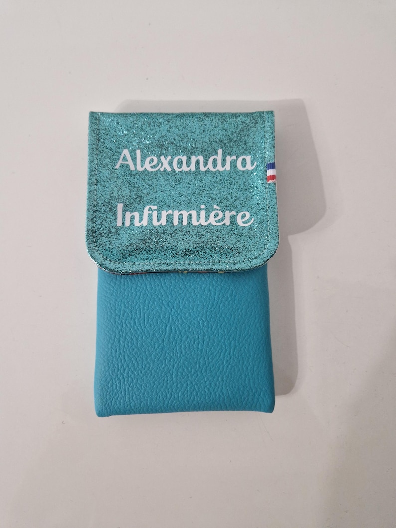 May include: A turquoise blue pouch with a glittery flap. The flap has the name "Alexandra" and the word "Infirmi&egrave;re" printed in white. A small French flag is on the right side of the flap.