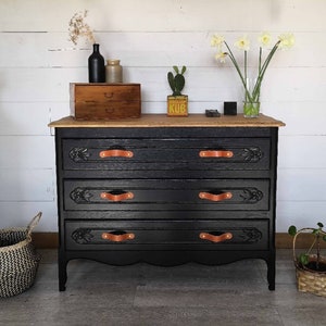 May include: A black dresser with four drawers and leather pulls. The dresser has a wood top and is sitting on a wooden floor. The dresser is decorated with a small wooden box with the word "KUB" on it.