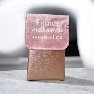 May include: A pink, glittery, and textured pouch with a flap. The flap has the name "Emma" and the text "Auxiliaire de pu&eacute;riculture" in white script. A small French flag is on the right side of the flap. The pouch is made of a metallic, rose-gold material.