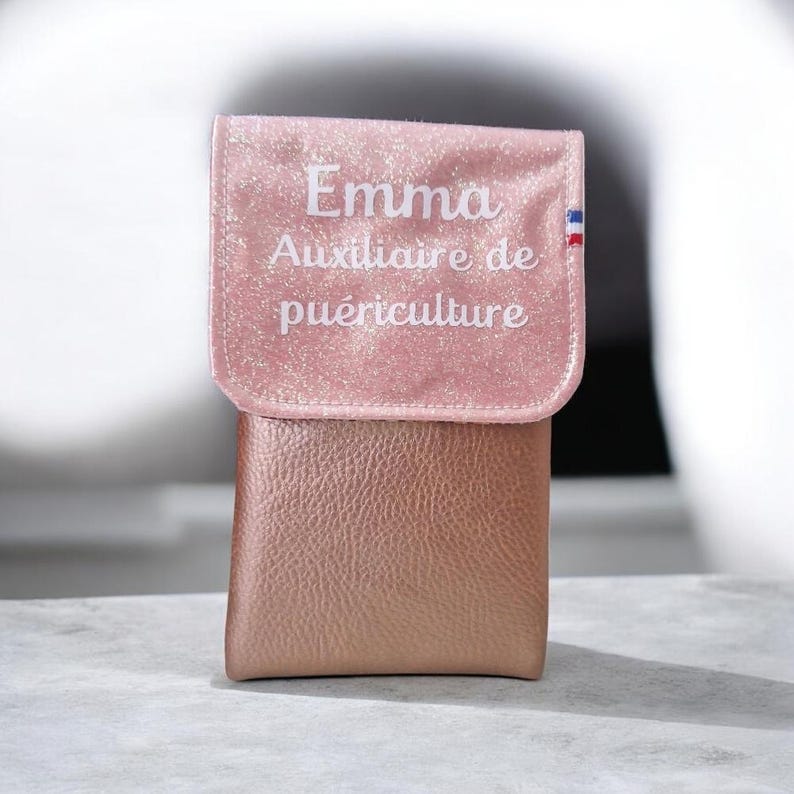 May include: A pink, glittery, and textured pouch with a flap. The flap has the name "Emma" and the text "Auxiliaire de pu&eacute;riculture" in white script. A small French flag is on the right side of the flap. The pouch is made of a metallic, rose-gold material.