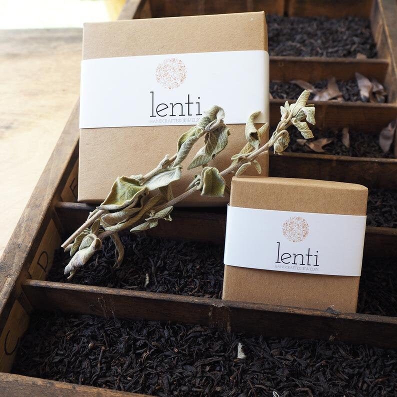 May include: Two brown boxes with white labels that say "lenti handcrafted jewelry" on them. The boxes are sitting on a wooden surface with dried leaves.
