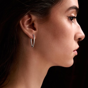 Puede incluir: Un par de pendientes de aro plateados en forma de V. Los pendientes tienen una superficie lisa y brillante. El diseño es minimalista y moderno. Se muestran en una oreja sobre un fondo oscuro. Un accesorio sencillo y elegante.
