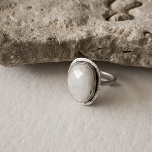 May include: A silver ring with a white moonstone set in a textured bezel. The ring is on a white background.