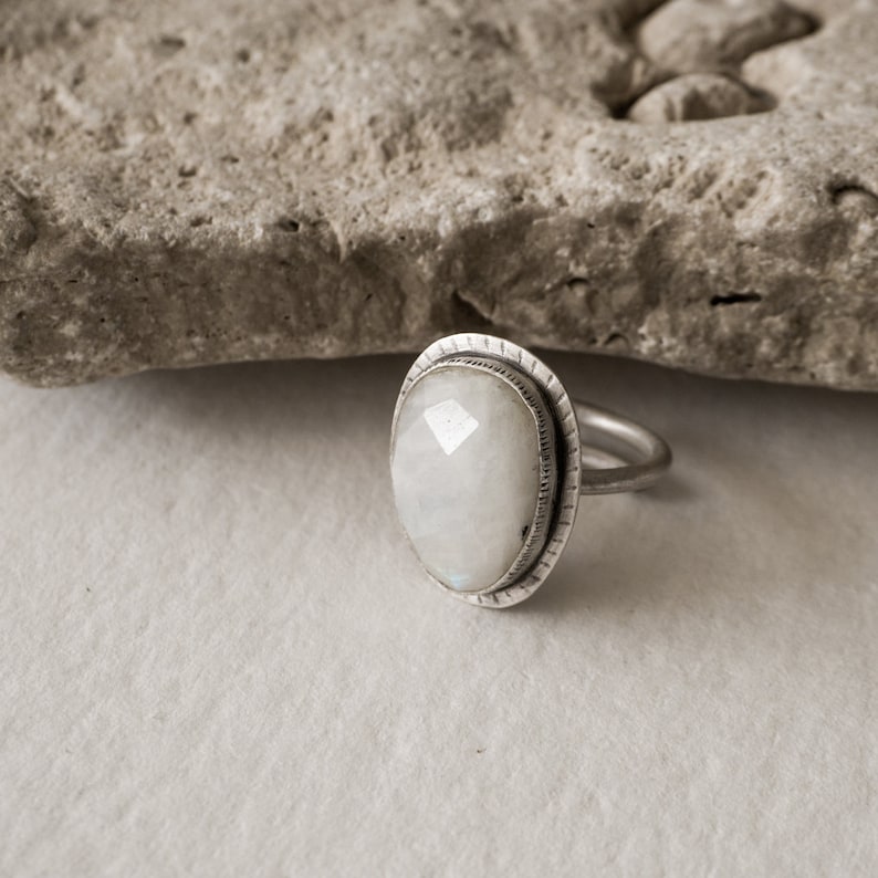 May include: A silver ring with a white moonstone set in a textured bezel. The ring is on a white background.