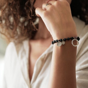Könnte beinhalten: Ein Armband aus schwarzem Lavastein mit drei silbernen Anhängern, darunter zwei runde Scheiben und ein offener Kreis.