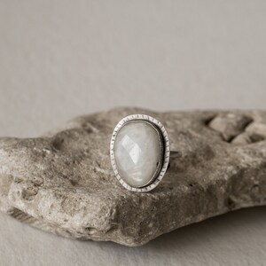 May include: A silver ring with a white moonstone set in a bezel setting. The ring is on a gray rock.