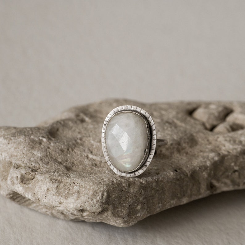May include: A silver ring with a white moonstone set in a bezel setting. The ring is on a gray rock.