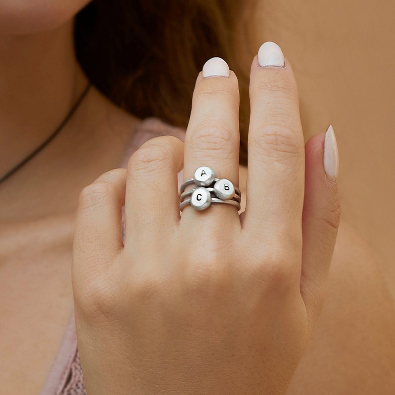 May include: A silver ring with three stacked bands, each featuring a small round charm with a letter on it. The letters are A, B, and C.
