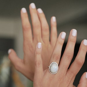 May include: A silver ring with a large oval moonstone set in a bezel setting. The ring is on a person's finger.