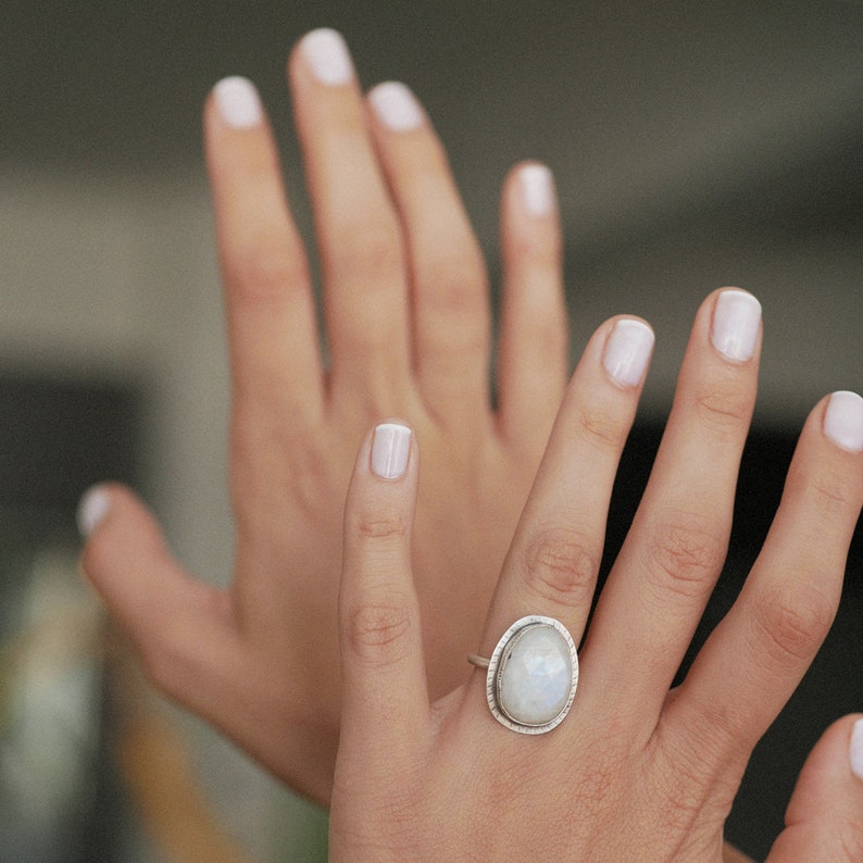 May include: A silver ring with a large oval moonstone set in a bezel setting. The ring is on a person's finger.
