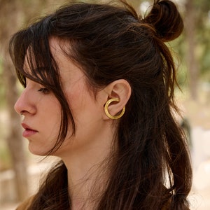 Puede incluir: Una mujer con el pelo largo y castaño lleva un pendiente de aro de oro. El pendiente de aro es de forma circular simple con una superficie texturizada.