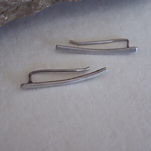 May include: A pair of silver ear cuffs, each with a curved, flat bar design. The cuffs are simple and minimalist.