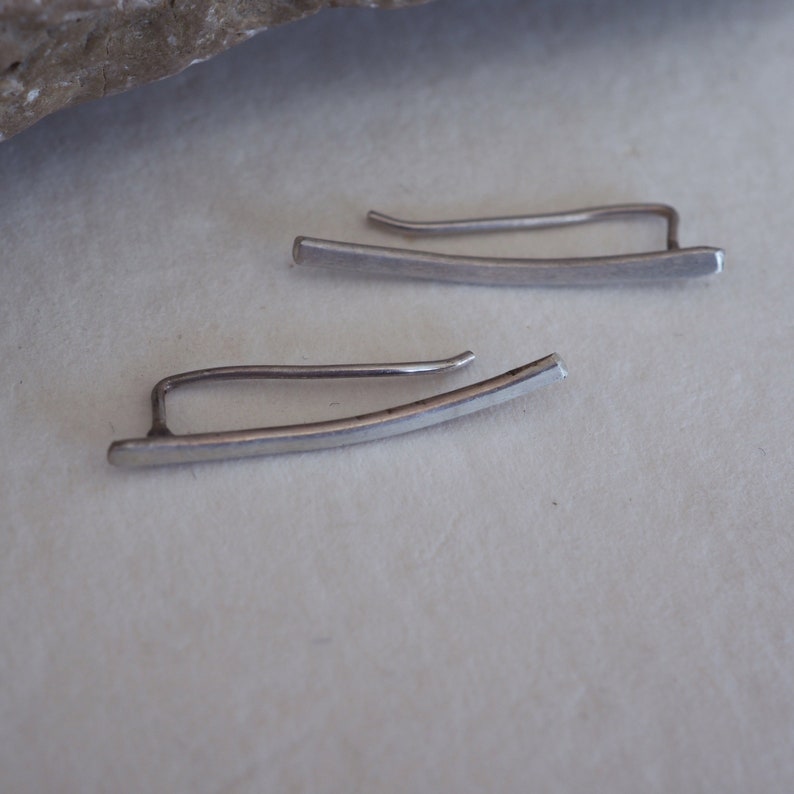 May include: A pair of silver ear cuffs, each with a curved, flat bar design. The cuffs are simple and minimalist.