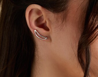 May include: A silver ear climber earring is shown on an ear. The earring has a curved, open design that follows the curve of the ear. The earring is made of a shiny, reflective metal.