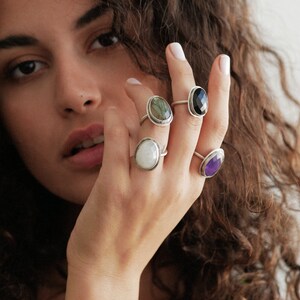 May include: A hand wearing four silver rings with oval-shaped gemstones. The rings feature a green, black, white, and purple stone.
